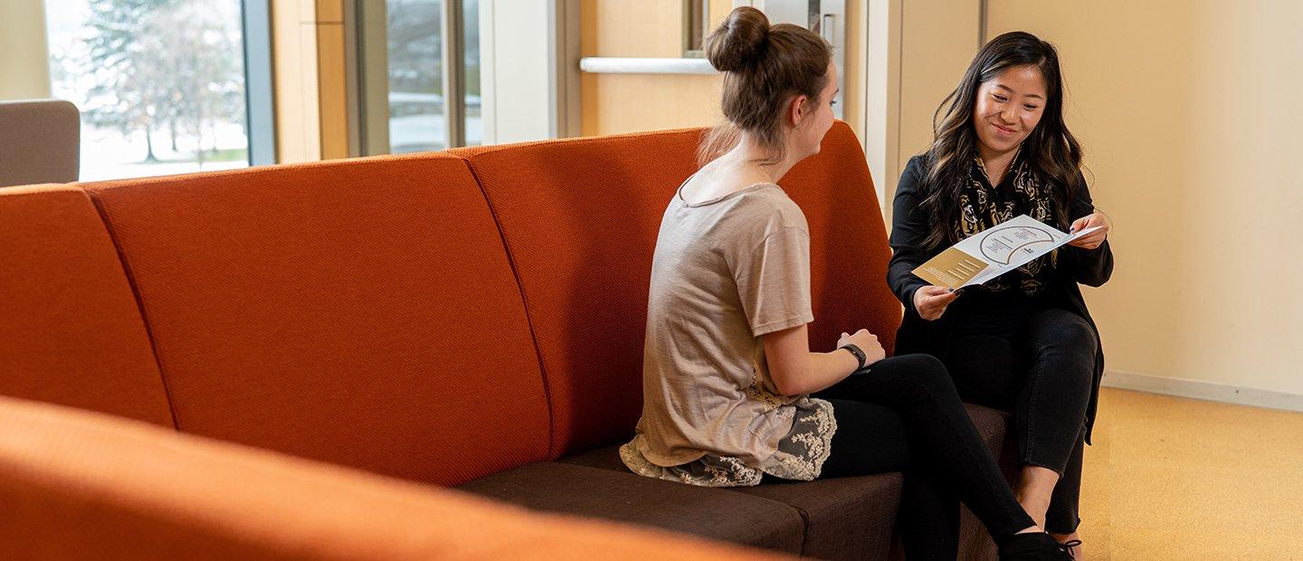 An adviser and a student seated on a couch, reviewing a brochure.